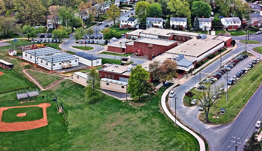 Aerial photograph of Bucknell Elementary School taken on April 28, 2015, prior to the start of our school's third renovation. The school is pictured from the northwest from a vantage point above University Drive. The hillside next to the school is intact. The gymnasium and subsequent classroom additions are visible. There are several mobile classroom trailers behind the school. 