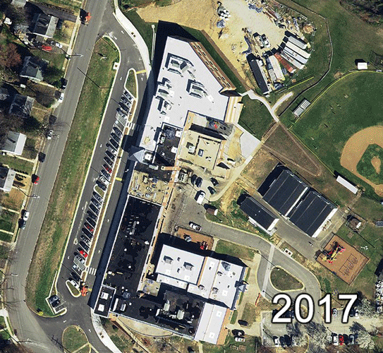 This animated series of aerial photographs shows Bucknell Elementary School from directly overhead. The images behind in 2017 and progress back in time to 2009, 1990, 1976, 1960, and 1937. In 1937, the future site of the school is an open field with trees to the north and a small dirt road running through the middle of the property. In 1960 the building appears as originally constructed. The building is similar in shape to the letter L with a long classroom wing running north to south, and the smaller nine-classroom addition running east to west. In 1976, additional classrooms have been added to the rear of the building and blacktops have been paved behind the school. In 1990 the gymnasium addition is visible, connected to the nine-classroom addition. In 2009, the new library has been added on the south side of the building. It juts out from the structure in an arc. The building has been re-roofed and two mobile classroom trailers are visible on the north side of the building near the ball fields. A playground has been added east of the blacktop behind the school. The 2017 photograph shows the renovation still in progress. Portions of the building are being re-roofed. Construction trailers and classroom trailers are visible around the school on the north and east. The classroom trailers have been set up on the blacktop.    