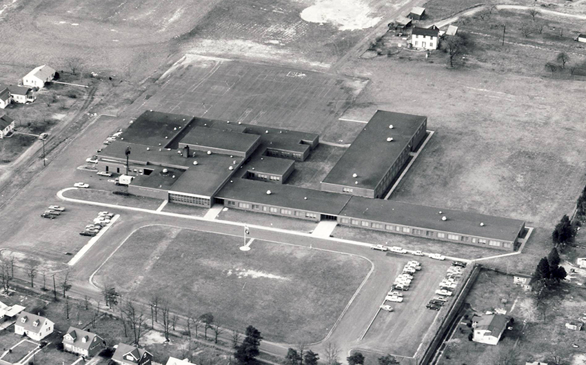 Black and white aerial photograph of Bryant Intermediate School taken in the 1960s. The building is a small structure with one and two-story classroom wings. Farm fields and a farmhouse are visible behind the school. Surrounding the school on the Quander Road side of the structure are new housing subdivisions.