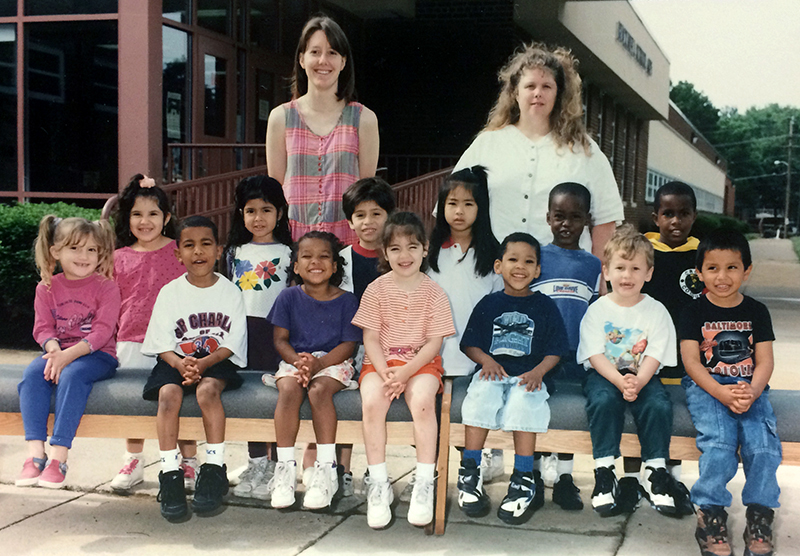 Color photograph of one of Bucknell's Head Start classes taken during the 1995 to 1996 school year. 13 children and two teachers are pictured. They are posed on the sidewalk in front of the main entrance to the building. 
