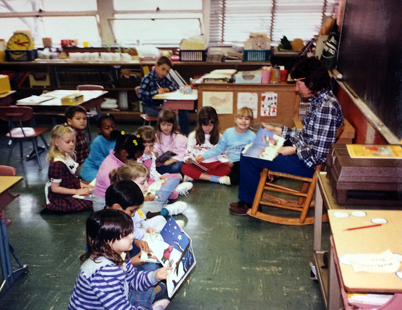 Color photograph of a classroom in 1989 with a lesson in progress. The teacher is seated in a rocking chair in front of the chalkboard. She holds a picture book in her lap. 12 children are pictured seated on a green tile floor. Nearly all of the children appear to be holding the same picture book as the teacher. The asbestos-laden green tiles, installed when the school first opened, have since been removed.