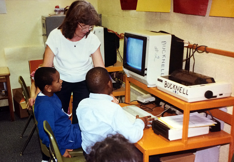 Photograph of two students working with a teacher at a computer workstation in 1989. A small CRT monitor, keyboard, and dot matrix printer are visible on the desk. 