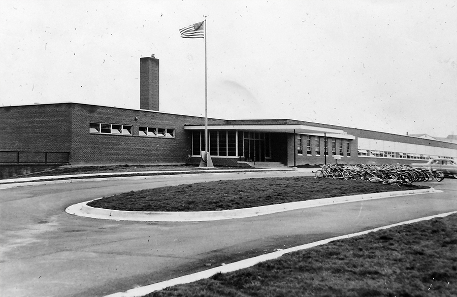 Black and white photograph of the front exterior of Bucknell Elementary School. The main entrance, cafetorium wing, and classroom wing are visible. Rows and rows of bicycles are parked on the grass and parking lot in front of the school. One car is visible, parked in the lot next to the bikes. 