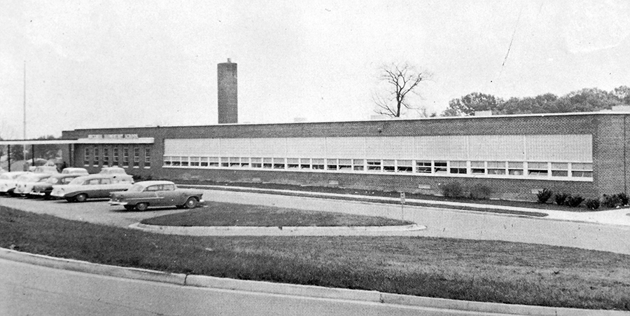 Black and white photograph of the front exterior of Bucknell Elementary School. The main entrance, classroom wing, and front parking lot are visible. 1940s and 1950s era cars are parked in the lot. From this vantage point, only the top-most story of the building is visible. The classroom wing has a large bank of windows that runs from one end to the other. 