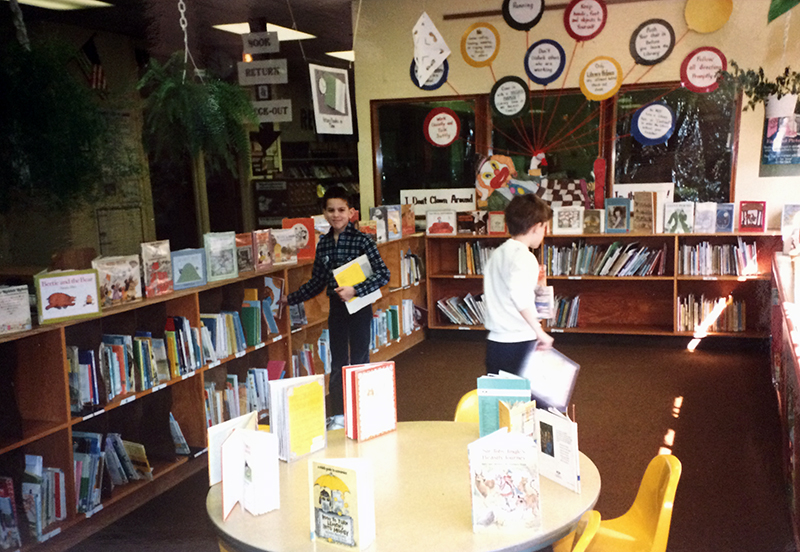 Photograph of two students in the library re-shelving books. The bookshelves are only two-rows tall and are lined with open books on top. A small circular table with yellow chairs is in the foreground. An office is visible in the background.  