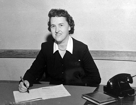 Black and white photograph of Bucknell's first principal, Sylvia D. Allen, taken in 1957. She is seated at her desk signing paperwork. A rotary-dial phone and notebook are on the desk to her left. 