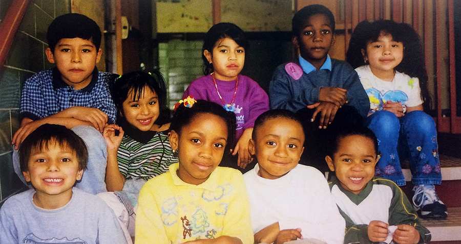 Photograph of nine students seated featured in our 2001 to 2002 yearbook. These students were new to Bucknell Elementary School that year and are of various ages. They are seated on a staircase inside the school with a hallway visible behind them in the distance.